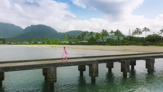 Aerial slow motion elegant lady in waiving dress walking by beach pier with cinematic mountain landscape on motion background. Happiness, freedom, dream trip, summer vacation concept. Hawaii nature US