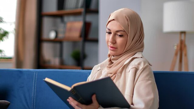 Muslim woman in hijab headscarf reading vintage religious book literature hobby at home couch