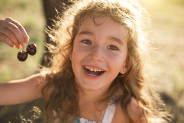 A cheerful sweet girl with cherry berries in her mouth. Funny summer portrait of a child with a...