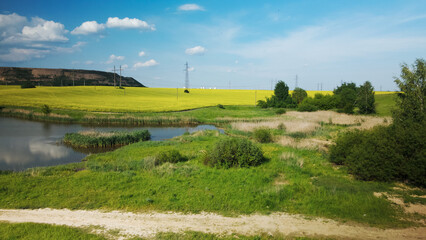 Blooming rapeseed field. On the edge of the field there is a pond. A mothballed city dump is visible on the horizon.Aerial photography.