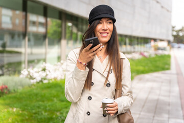 Businesswoman with a coffee in her hand and sending a voice note with the phone, wearing a trench coat