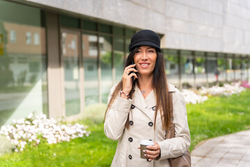 Businesswoman with a coffee in her hand and talking on the phone outside the office, wearing a trench coat