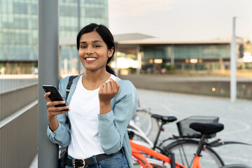Attractive smiling Indian woman holding mobile phone, shopping online looking at camera outdoors....