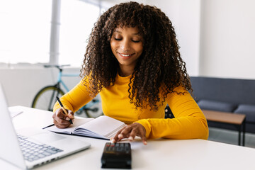 Smiling young african american woman working with laptop computer at home office - Female entrepreneur using calculator while taking notes at workplace - obrazy, fototapety, plakaty