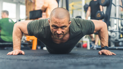 Bald caucasian tired manwearing a green t-shirt trying hard to do a push-up on gym flooring, looking directly at a camera. High quality photo