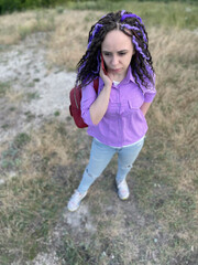 Young beautiful woman talking on mobile phone, standing in hilly terrain. Cute vivid female with dreadlocks speaking on smartphone, standing outdoors in cloudy, windy, summer weather.