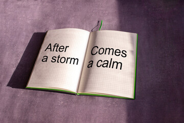 An open notebook is on the gray background. The words "after storm comes a calm" is written on a blank sheet of notebook. Learning, education concept.