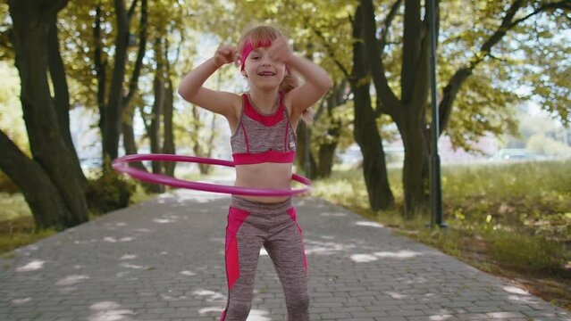 Athletic fitness toddler girl training playing twisting Hula hoop circle ring around waist. Workout gymnastics in city park at morning. Child kid enjoying healthy lifestyle. Active children motivation