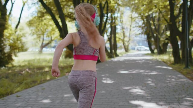 Back view of athletic fitness sport runner toddler girl training marathon run, listening music in headphones. Workout cardio outdoor in park. Child kid enjoy healthy lifestyle. Active children jogger