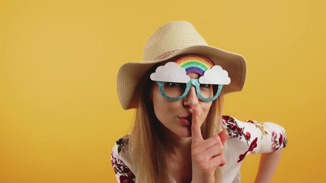 Funny carefree caucasian young adult girl in glittered rainbow glasses shushing the camera with index finger and nodding her head. High quality 4k footage