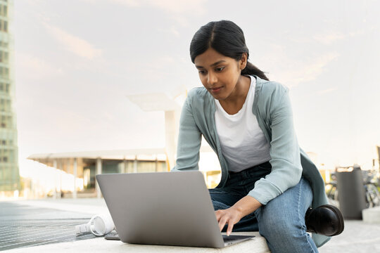 Pensive Indian student using laptop computer studying, exam preparation sitting in modern university campus, online education. Young asian copywriter typing on keyboard working freelance project 