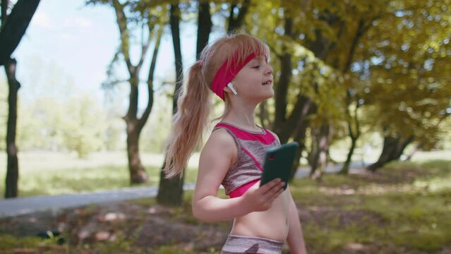 Athletic fitness sport runner child girl walking and using smartphone listening music with wireless earphones. Workout cardio in morning park. Children toddler kid jogger enjoying healthy lifestyle