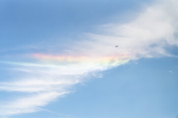 Naklejka na ściany i meble Beautiful sky background photograph of an airplane flying past a partial rainbow reflecting on the white clouds in the sky on a sunny day.