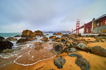 Stunning sandy beach waves crashing into rocks covered in mussels by Golden Gate Bridge on foggy...