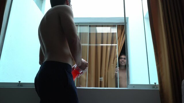 two shirtless friends chatting by the window of two hotel rooms together in the morning after a night out