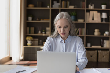 Serious middle-aged businesslady work on laptop seated at desk staring at screen read e-mail,...