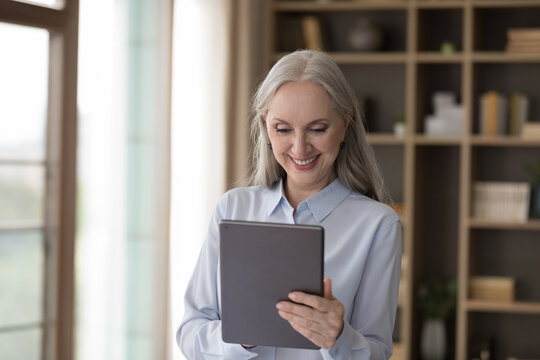 Smiling attractive middle-aged businesslady stand at workplace hold digital tablet working on-line, make task, learn new software, buy services on internet. Modern tech, business application concept