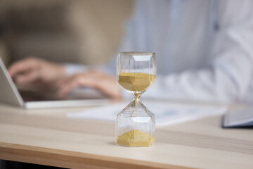 Unknown businesswoman sit at desk working on laptop, focus on hourglass showing passage of workday time. Deadline, time is money, billable hours, productive and effective work day, workflow concept