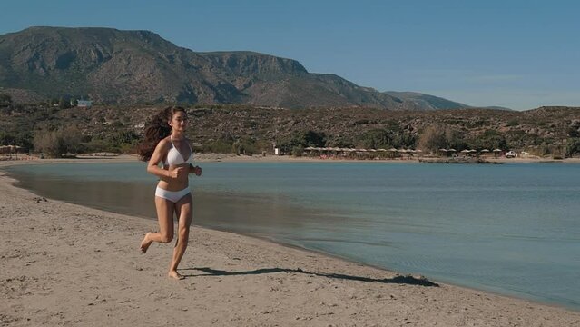 Young Asian women exercising with outdoor running mountain sea background. Exotic Woman in white Bikini Run to Ocean in beautiful sea water. Concept of vacation, Tourism, healthy running, exercise.