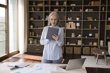 Confident attractive older businesswoman stand at workplace hold digital tablet make online task,...