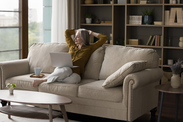 Serene middle-aged attractive woman resting on cozy sofa with teacup and laptop looks peaceful relaxing at home with eyes closed, spend carefree weekend at modern home. Technologies, leisure concept
