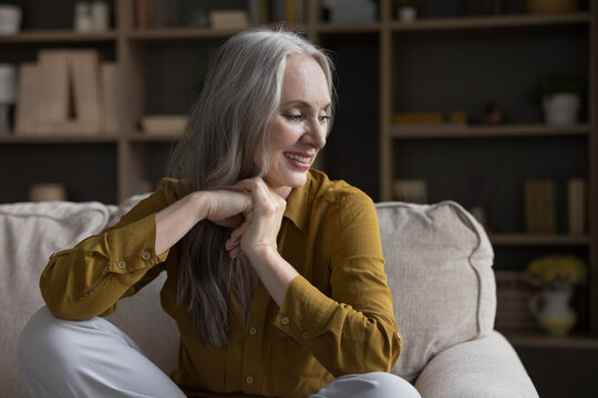 Calm beautiful middle-aged single woman sitting on cozy sofa, smiling staring aside looks optimistic and carefree enjoy rest alone in fashionable living room at modern own home. Relax, leisure concept