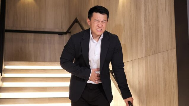 Stressed overworked asian businessman feeling pain stomach ache. A young worker stands on the stairs and holds stomach, excited about the loss of business, work, failed. The nervous system is failing.