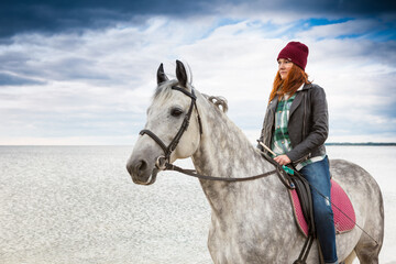 wearing jeans, jacket and fall hat horsewoman rides astraddle along sea shore