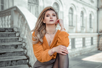 A middle-aged woman looks like a good blonde with curly beautiful hair and makeup on the background of the building. She is wearing a yellow sweater.