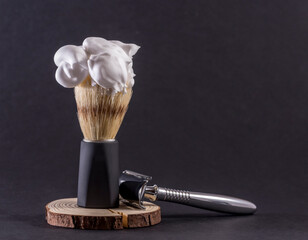 boar bristle shaving brush with shaving foam on top and with a black handle next to a shaving t-shaped shaving machine on a black background