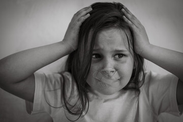 Black and white portrait of little afraid panicked girl with long dark hair grabbing head. Mouth sealed with medical plasters. Protest against domestic violence, abuse, fighting, conflicts, bullying.