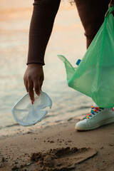 Cropped photo of African woman collecting spilled trash garbage from sand on beach in green plastic bag. Womans hand picking up white used plastic bowl container. Ecology, pollution, earth day. 