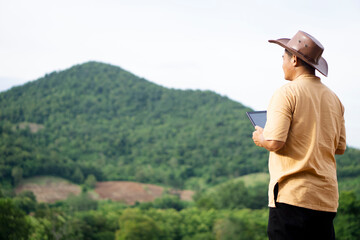 Side view of Asian man botanist wears hat, holds smart tablet, stand at nature forest, look forward to mountain. Concept : Nature survey, use wireless technology and internet to manager environment.