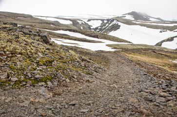 Winding track with ravel and rocks, partly covered with snow in a mountain landscape near...