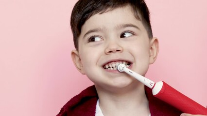 cute little boy using electric toothbrush for daily teeth cleaning.kid with dark red towel on neck.pink background,4k close up.dental care,dentist for kids concept. smiling boy, diastema,space between - Powered by Adobe
