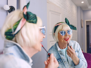 Smiling elderly senior stylish woman in blue sunglasses and denim jacket using red lipstick by the mirror in stylish loft interior. Style, fashion, make up, anti age concept