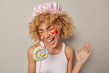 Cheerful carefree young woman with curly hair holds round colorful lollipop keeps palm raised sings...