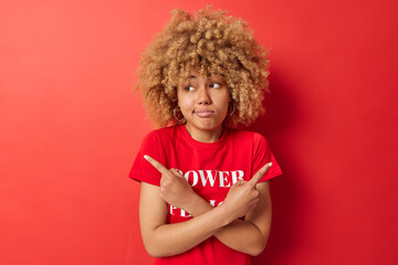 Confused curly haired young European woman points sideways chooses between two options has hesitant expression dressed in casual t shirt isolated over red background. People and choice concept