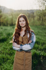 A farmer woman in her work clothes, plaid shirt and apron, stands in the field on the green grass and smiles in the setting sun