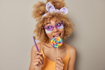 Photo of lovely woman with curly bushy hair wears purple sunglasses headband and orange t shirt licks multicolored lollipop applies beauty patches holds toothbrush isolated over grey background.