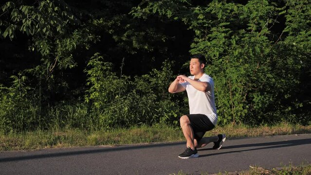 Full length shot of active fit young korean man athlete doing lunges during workout at running track sunset background of green trees. Summer time male workout spot outdoor. Sport healthy concept.