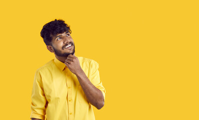 Obraz premium Cheerful stylish Indian guy pensively looks at copy space touching his chin, isolated on orange background. Handsome young ethnic bearded man in yellow shirt has plan or idea in mind. Web banner.