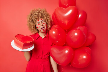 Bad mood on holiday. Frustrated curly haired European woman holds heart shaped balloons and delicious cake expresses negative emotions isolated over vivid red background. Party time concept.