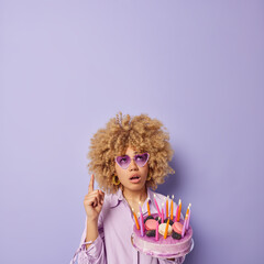 Vertical shot of surprised woman with curly hair points index fingr overhead has wondered expression holds tasty cake with burning candles trendy sunglasses and shirt isolated over purple wall