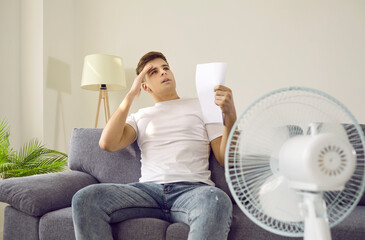 Summer heat. Sweaty man feels bad because of terrible heat sitting at home in front of fan during...