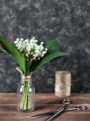 Bouquet of lilies of the valley in vintage glass vase with old scissors and skein of twine on wooden table on black concrete background with copy space. Rustic still life with spring flowers.