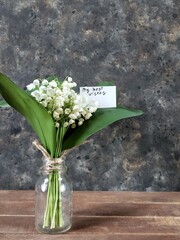 Bouquet of lilies of the valley in vase with white greeting card with inscription My best wishes on wooden table on black concrete background with copy space. Rustic still life with spring flowers.
