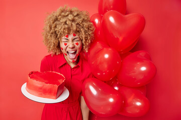 Emotional curly haired woman exclaims loudly from joy wears festive dress holds heart shaped cake and bunch of inflated balloons poses against vivid red background. Festive occasion concept.