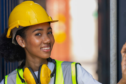 Young African American Trainee In Safety Suit, Yellow Hard Hat And Earmuffmuff Stand Smiling Next To Big Container In A Shipyard. African Curly Hair Female Worker In Smile. Female Loader With Earmuff