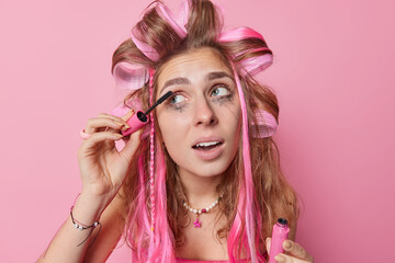 Headshot of long haired young European woman applies hair rollers and mascara looks away undergoes beauty treatments prepares for first date wants to look beautiful isolated over pink background.